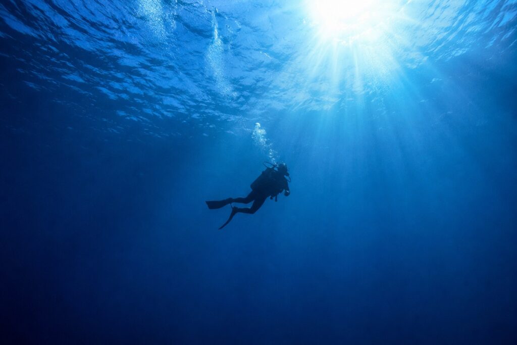 Scuba diver hovering alone in open blue water with sunlight rays from above, representing individual diver competence over agency brand loyalty